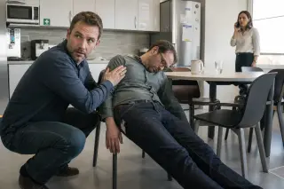 Office kitchen scene during lunch with one man supporting a collapsed colleague and another co-worker calling for help, natural light, subtle and authentic atmosphere.